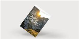Ein Buch über das Kaisergebirge mit majestätischen Bergen im Hintergrund. Der Sonnenuntergang verleiht der Szene eine warme Atmosphäre.