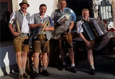 Vier Männer in traditionellen bayerischen Trachten stehen lächelnd zusammen. Sie halten Musikinstrumente wie eine Tuba, ein Akkordeon und Trompeten.