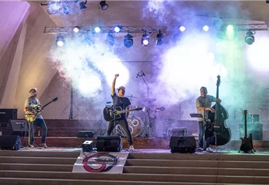 Eine Band spielt auf einer Bühne mit bunten Lichtern und Nebel. Der Sänger hebt die Hand, während die Musiker mit ihren Instrumenten auftreten.