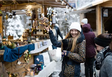 A cheerful young woman at a Christmas market holds a decorative glass lantern. In the background, there are stalls with festive lighting and other visitors.
