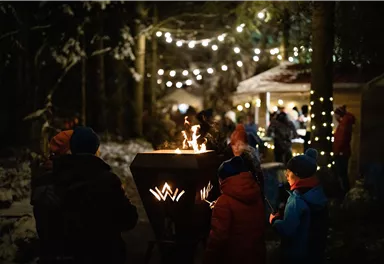 A winter scene with a fire pit surrounded by people. In the background, there are fairy lights creating a cozy atmosphere.
