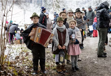 A group of children in traditional clothing stands on a path in the snow. Some of them are holding musical instruments and smiling at the camera.