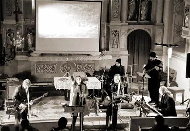 A band performs in a church with a projector in the background. The musicians play various instruments and stand on a stage.