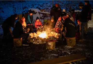 A group of people sits around a campfire in a wintery setting. The atmosphere is cozy and inviting.