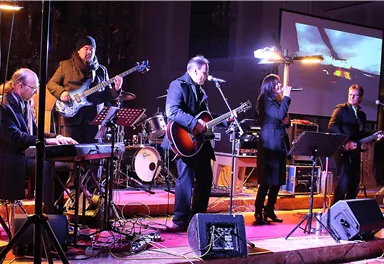 A band is playing live on a stage with various musical instruments. In the background, lights and a screen can be seen.
