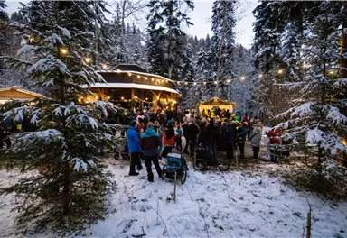 A winter celebration in the forest with many people. The surroundings are covered in snow, and the light of the event creates a cozy atmosphere.