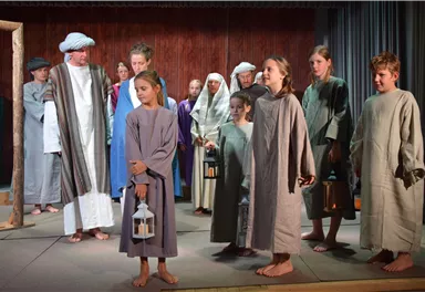 A group of children and adults in historical costumes stands on a stage. They are holding lanterns and are attentively looking towards the audience.