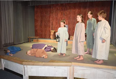 A theater performance with several children in historical costumes. Some are standing while others are lying on the stage.