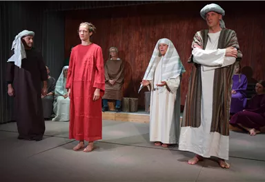 A theatrical performance featuring actors in historical costumes. The scene appears dramatic, with various characters on stage.