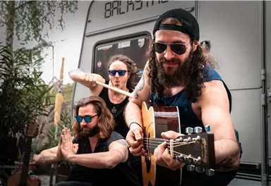 Drei Musiker spielen in einem entspannten Ambiente. Im Hintergrund sind Pflanzen und ein Wohnwagen zu sehen.