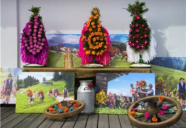 Traditionelle Blumenkränze in bunten Farben stehen auf einem Tisch. Im Hintergrund sind Bilder von der Alm und fröhlichen Menschen mit Kühen zu sehen.