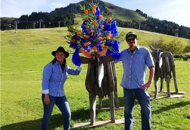 Ein Paar steht neben einer Skulptur einer Kuh, die mit bunten Blumen geschmückt ist. Im Hintergrund sind sanfte Hügel und eine klare Himmel zu sehen.