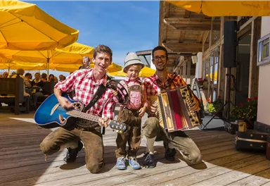 Two musicians in traditional clothing are playing with a child. They are standing on a terrace with yellow umbrellas in the background.