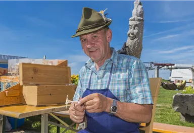 A smiling man is wearing a traditional hat and working at a table outdoors. Behind him, wooden crates and a green meadow can be seen.