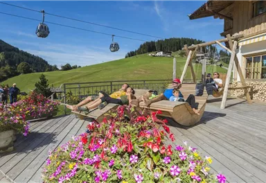 Eine entspannte Terrasse mit Holzschaukeln und bunten Blumenbeeten. Im Hintergrund sind grüne Hügel und Seilbahnen zu sehen.