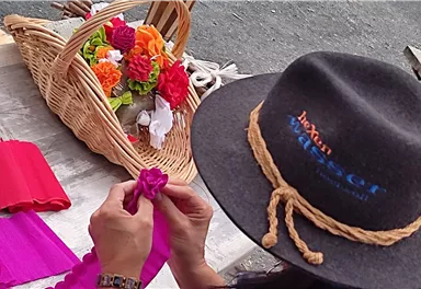 Eine Person bastelt Blumen aus buntem Stoff. Im Hintergrund ist ein Korb mit bereits fertiggestellten Blumen zu sehen.