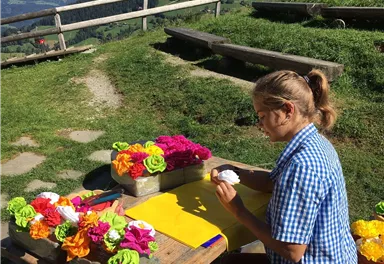 Eine Person sitzt an einem Tisch und bastelt farbenfrohe Blumen aus Papier. Im Hintergrund sind grüne Wiesen und Berge zu sehen.