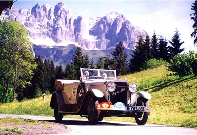 Ein klassisches Auto fährt eine malerische Straße in den Bergen. Die Umgebung ist grün mit hohen Bäumen und beeindruckenden Berggipfeln im Hintergrund.