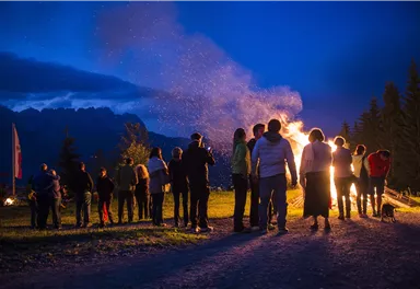 A group of people is standing around a campfire in the dusk. In the background, trees and mountains can be seen.