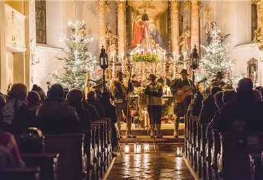 A festively decorated church with Christmas trees and candlelight. A music group plays in front of the audience in the beautiful surroundings.