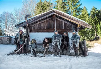 A group of people in traditional costumes stands in front of an old wooden house in the snow. In the background, there are trees and a blue sky.