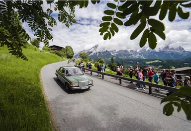 A group of people is hiking on a path through a green landscape. In the background, gentle hills and a small village are visible.