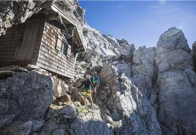 A mountain hut stands between rocky cliffs. Two hikers explore the surroundings under a clear blue sky.