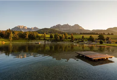 Ein ruhiger See mit einem Holzsteg und Bergen im Hintergrund. Die Landschaft ist grün und die Atmosphäre ist friedlich.