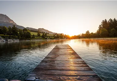 Ein ruhiger See mit einem Holzsteg, der ins Wasser führt. Im Hintergrund scheinen die Berge und die Sonne aufgeht in der Morgenluft.