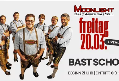 A group of men in traditional Tyrolean attire stands together. In the background, there is a banner for an event with live music on Friday, the 20th of March.