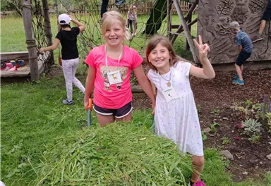 Zwei Mädchen stehen lächelnd vor einer Schubkarre mit frischem Gras. Im Hintergrund spielen andere Kinder auf einem Spielplatz.