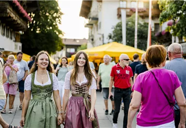 Zwei Frauen in traditionellen Trachten gehen fröhlich durch eine belebte Straße. Im Hintergrund sind Menschen und gelbe Pavillons zu sehen.