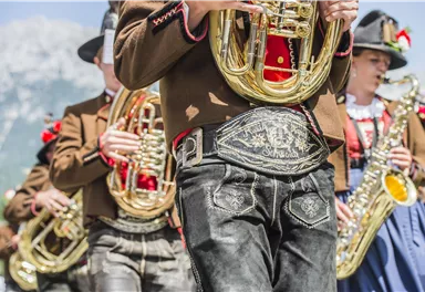 Eine Gruppe von Musikern in traditionellen Kleidern spielt Blasinstrumente. Im Hintergrund sind Berge und eine sonnige Landschaft zu sehen.