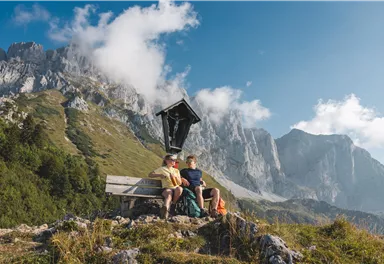 Two people are sitting on a bench in the mountain landscape. In the background, high mountains and a clear sky are visible.