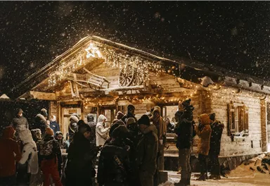 A cozy cabin at night, surrounded by heavy snowfall. Many people are standing outside, enjoying the festive lighting.