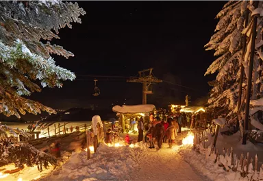 A snow-covered street at night, illuminated by torches and surrounded by snow-covered trees. In the background, people and a gondola lift can be seen.