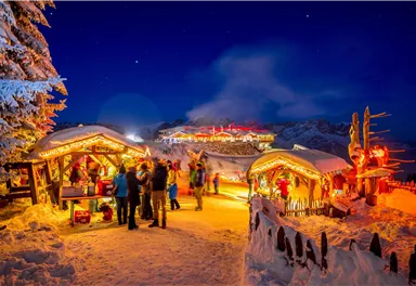 A festive Christmas market in a snowy landscape. The stalls are decorated with lights and people enjoy the atmosphere at night.