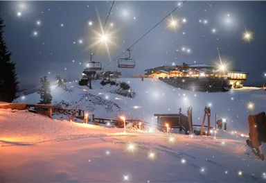 A snowy mountain area at night with a ski lift and a building in the background. The sky is adorned with sparkling elements that create a magical atmosphere.