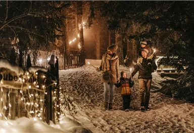 A family is walking at night on a snowy path, surrounded by festive lighting. The atmosphere is cozy and wintry.