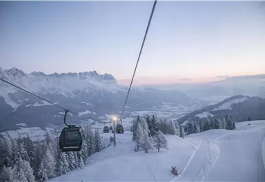 A picturesque winter landscape with snow-covered mountains and a cable car. The sky is clear with gentle colors of the sunset.