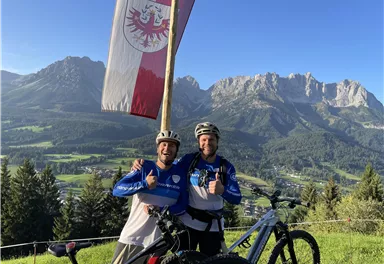 Two cyclists stand smiling in front of an impressive mountain backdrop. In the background, a flag is waving, and the landscape is green and picturesque.