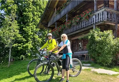 Two cyclists are standing in front of a traditional wooden house in nature. In the background, green trees and a beautiful view can be seen.