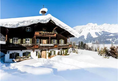 Ein schönes Chalet im Schnee mit einem schneebedeckten Dach. Im Hintergrund sind beeindruckende Berge zu sehen.
