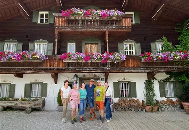 Eine Gruppe von sechs Personen steht vor einem traditionellen Holzhaus mit bunten Blumenkästen. Das Haus hat große, grüne Fensterläden und ist von einer schönen Landschaft umgeben.