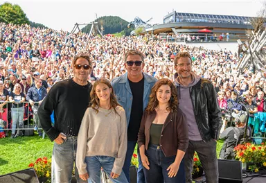 Fünf Personen stehen lächelnd vor einer großen Menschenmenge. Im Hintergrund sind Berge und ein Gebäude zu sehen.