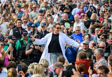 Ein Mann in einem weißen Laborkittel steht im Mittelpunkt einer großen Menschenmenge. Die Zuschauer applaudieren und nehmen Fotos auf.