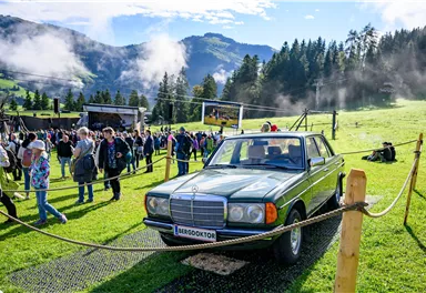 Ein klassisches Auto steht auf einem grünen Feld. Im Hintergrund sind Menschen und Berge zu sehen, und die Atmosphäre wirkt festlich.