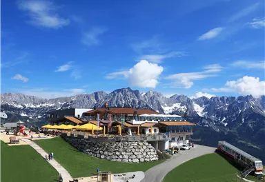 A picturesque mountain landscape with a restaurant and beach umbrellas. In the background, impressive mountains and a blue sky can be seen.