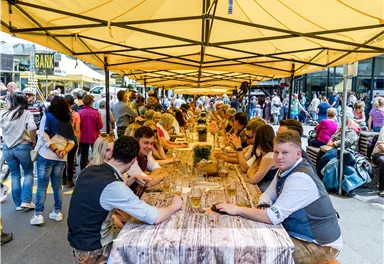 Eine belebte Straßenveranstaltung mit langen Tischen unter einem gelben Zelt. Menschen sitzen zusammen, genießen Essen und trinken, während eine große Menge im Hintergrund steht.