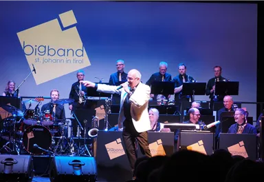 Ein Sänger tritt auf der Bühne auf, während eine Bigband hinter ihm spielt. Die Musiker sind in formellen Kleidern und konzentrieren sich auf ihre Instrumente.